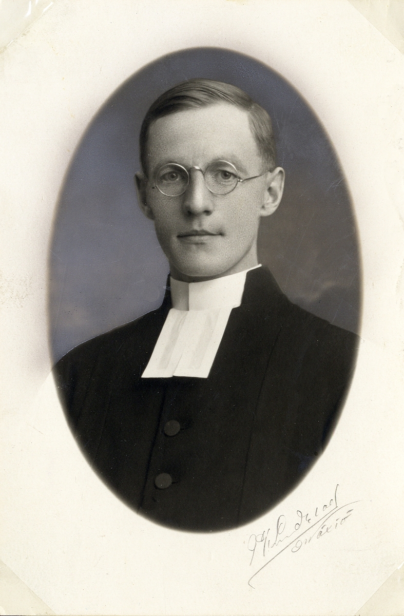 Foto av en man med glasögon, klädd i prästrock med prästkrage. 
Bröstbild, halvprofil. Ateljéfoto.
