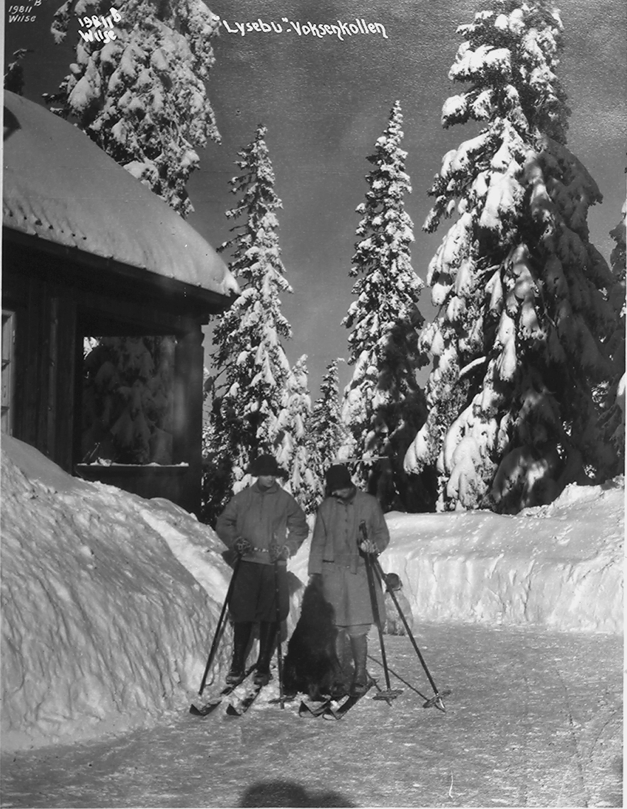 Kvinne og mann står med ski på bena foran Lysebu. Påskrevt bildet "Lysebu, Voksenkollen". Fotografert 1926.