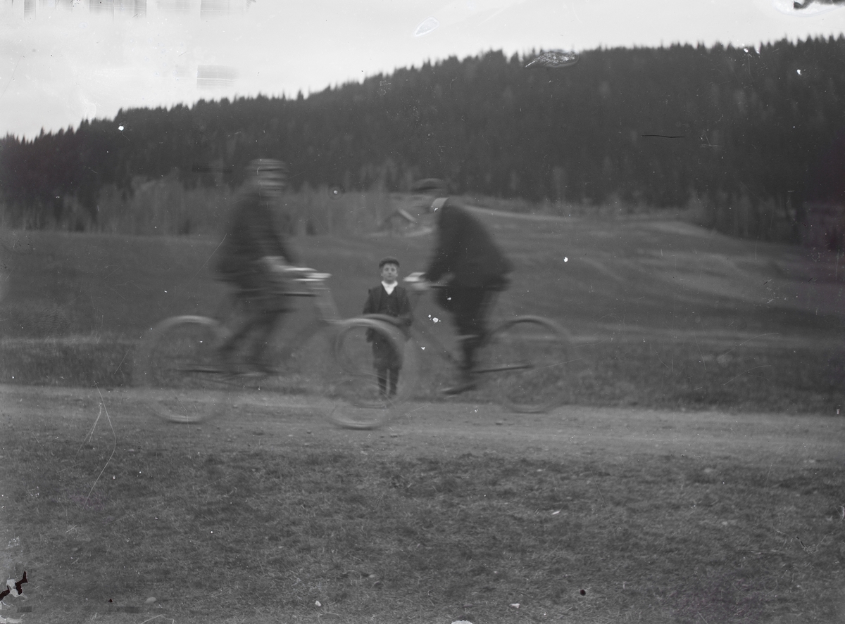 Motiv av to menn på hver sin sykkel som sykler forbi et barn som poserer for kamera.