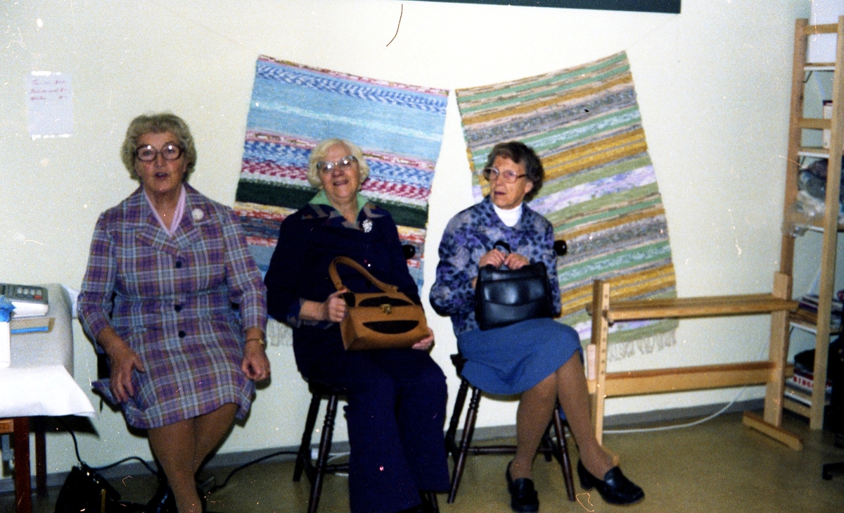 Julförsäljning av hantverk i pensionärernas hobbylokal på Våmmedalsvägen år 1977. Från vänster: Hulda Olsson, okänd kvinna samt Valborg Smitt (1901 - 1986). Bakom kvinnorna hänger två handvävda mattor på väggen.