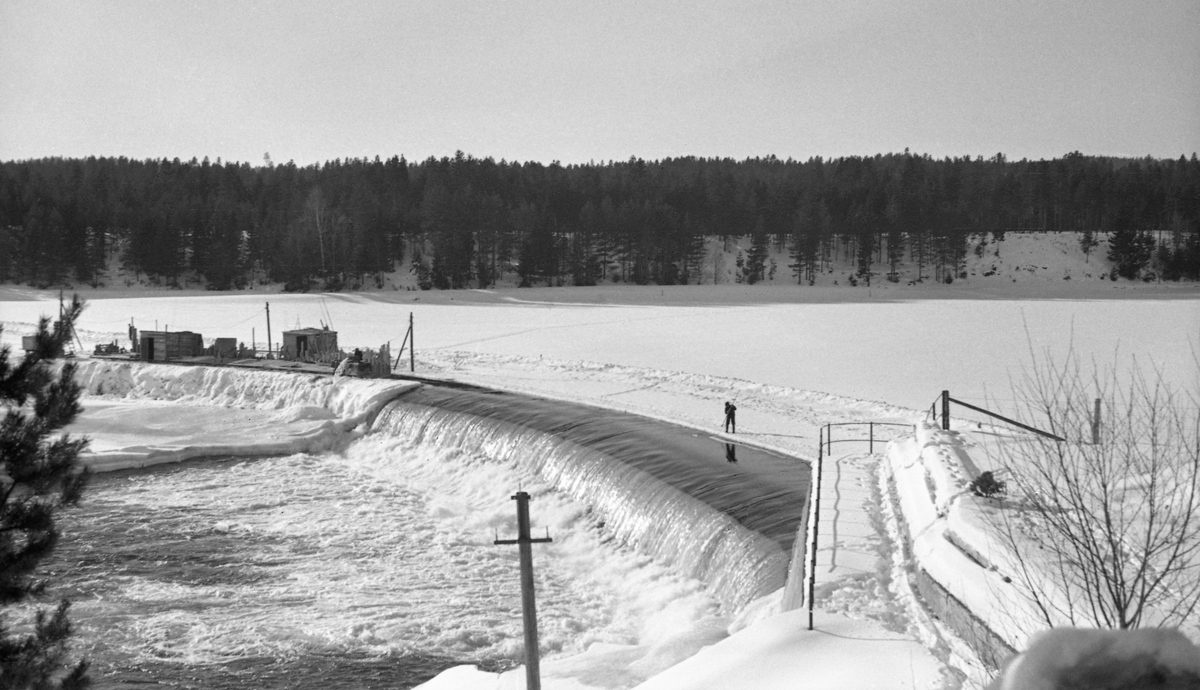 Kraftverksdammen ved Skjefstadfossen i Heradsbygda i Elverum.  Fotografiet er tatt i februar 1942, på et tidspunkt da bare den østre, djupeste og mest vannrike delen av elveleiet fra damkrona og nedover var åpent.  Den vestre delen av elveleiet derimot, var is- og snødekt.  Fotografen har stått på Glommas østre bredd med kameraet vendt mot den buete damterskelen.  I forgrunnen, til høyre, er det en murt "utstikker" med rekkverk, et slags utsiktspunkt mot dammen.  Om lag midt på dammen, til venstre i dette bildet, er det reist et par brakker, antakelig i samband med anleggsarbeid som ble gjort vinterstid, da det var liten vannføring.  Vi ser også at det er lagt strøm på midlertidig oppsatte stolper ut mot brakkene, antakelig for å få energi til belysning på arbeidsstedet i den mørke vintersesongen med korte dager. En mann sto på iskanten mot råka ved damkrona da fotografiet ble tatt.  I bakgrunnen, på vestre side av vassdraget, vokste det barskog. 