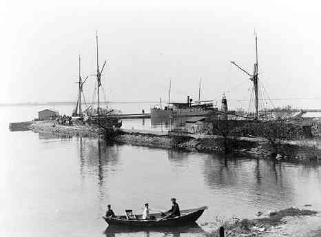 En man och hans två söner i en roddbåt söder om Gränna hamn, de har varit ute på fiske. I hamnen, till höger, ligger Motala Express och tre segelskutor vid kaj. På kajen ligger ett upplag med timmer.
