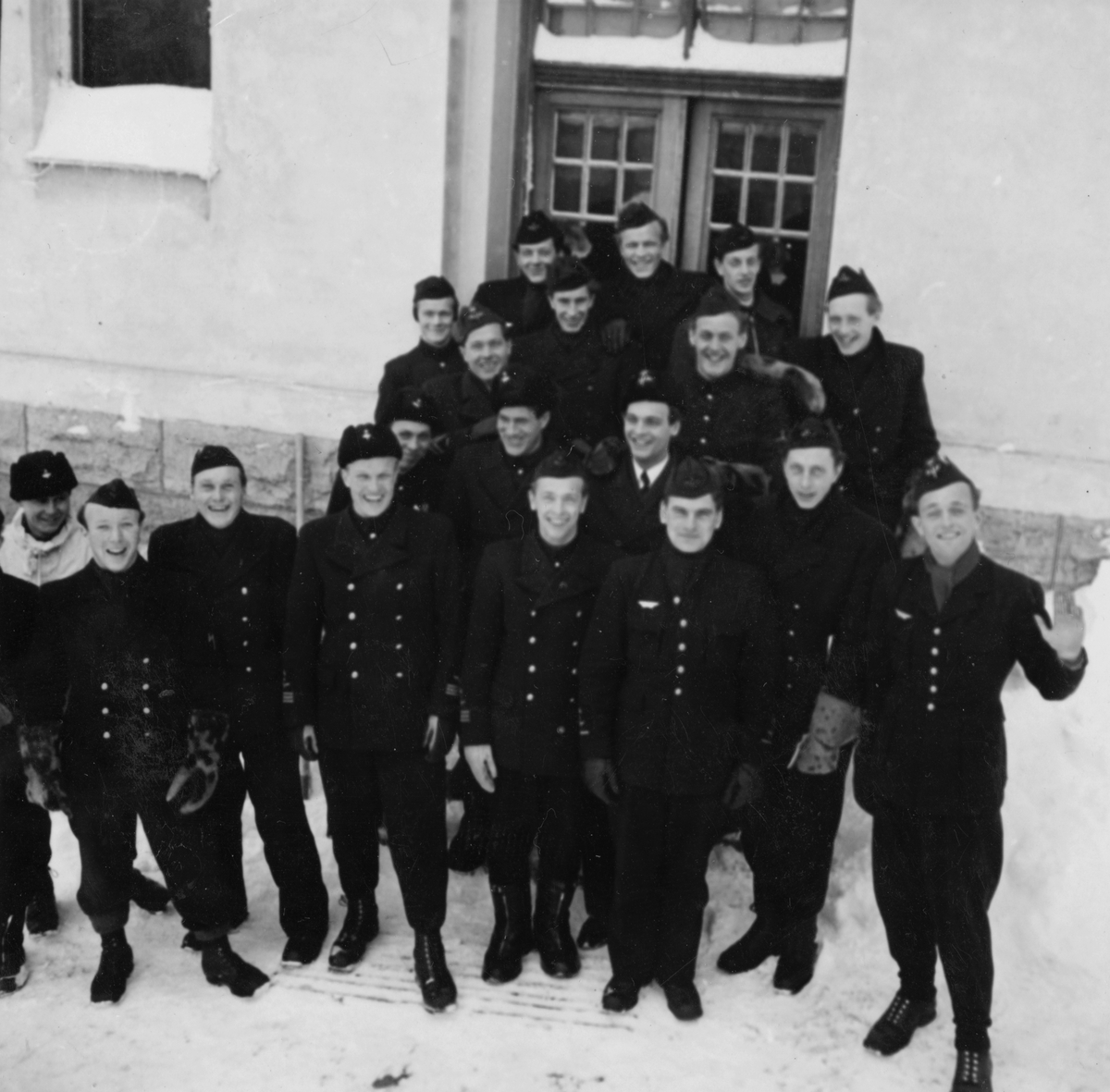 Gruppfoto av mekaniker och flygförare från F 14 före den planerade flygningen Halmstad-Kiruna i februari 1946. 19 män samlade framför hus. Flygningen slutade i nödlandning i Härnösandstrakten den 10 februari 1946.
