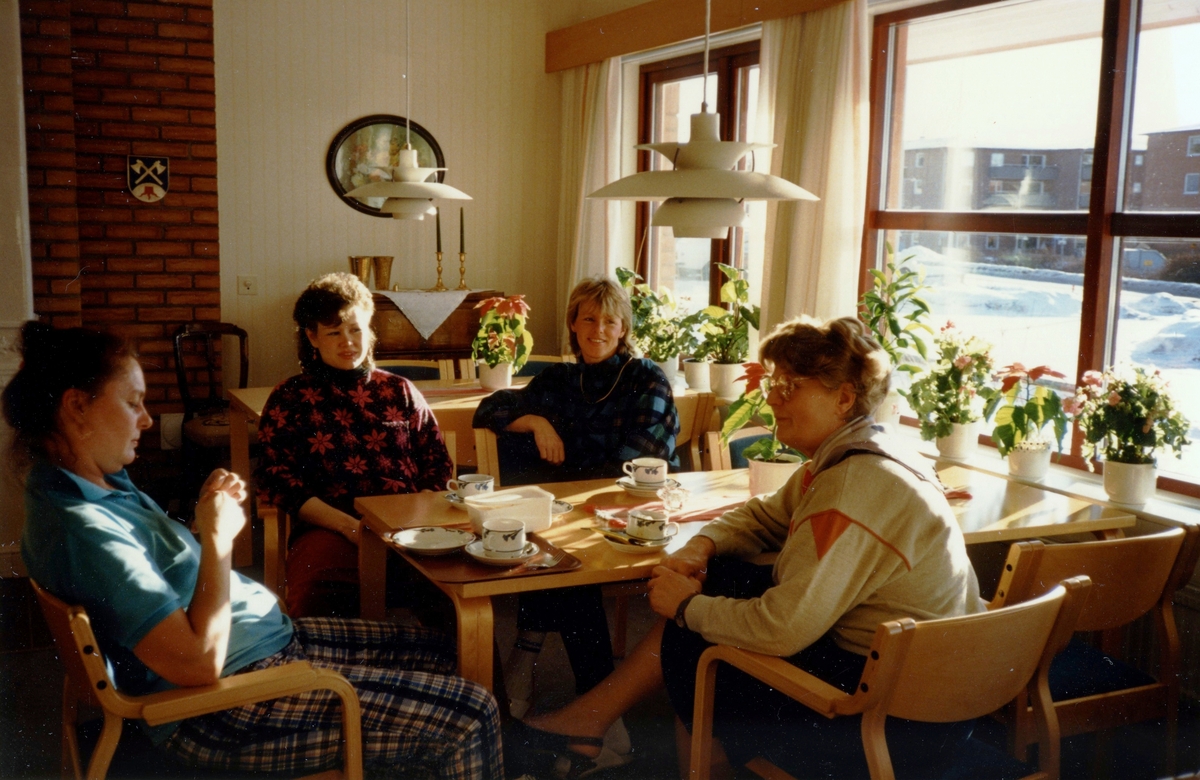 Fyra kvinnliga personal sitter och fikar runt ett bord på Brattåsgården (Streteredsvägen 5) cirka 1986 - 1990. Från vänster: 1. Okänd, 2. Eva Agfjord, 3. Lillvor Sjöström samt 4. föreståndare Aina Ekstedt. Till höger, genom fönstret, ses Vommedals lägenhetsområde.