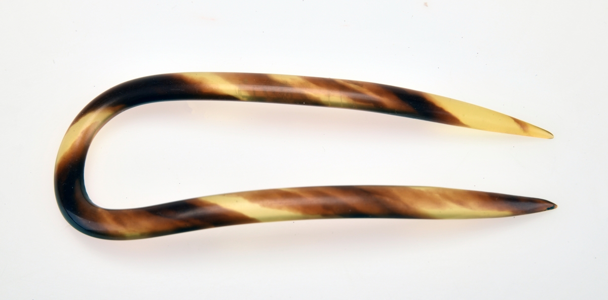 En hårkam laget av celloid i fargene brunt og gult (melert). Kammen er buet og har U-form. Den har to tenner som er runde og spisse i enden.