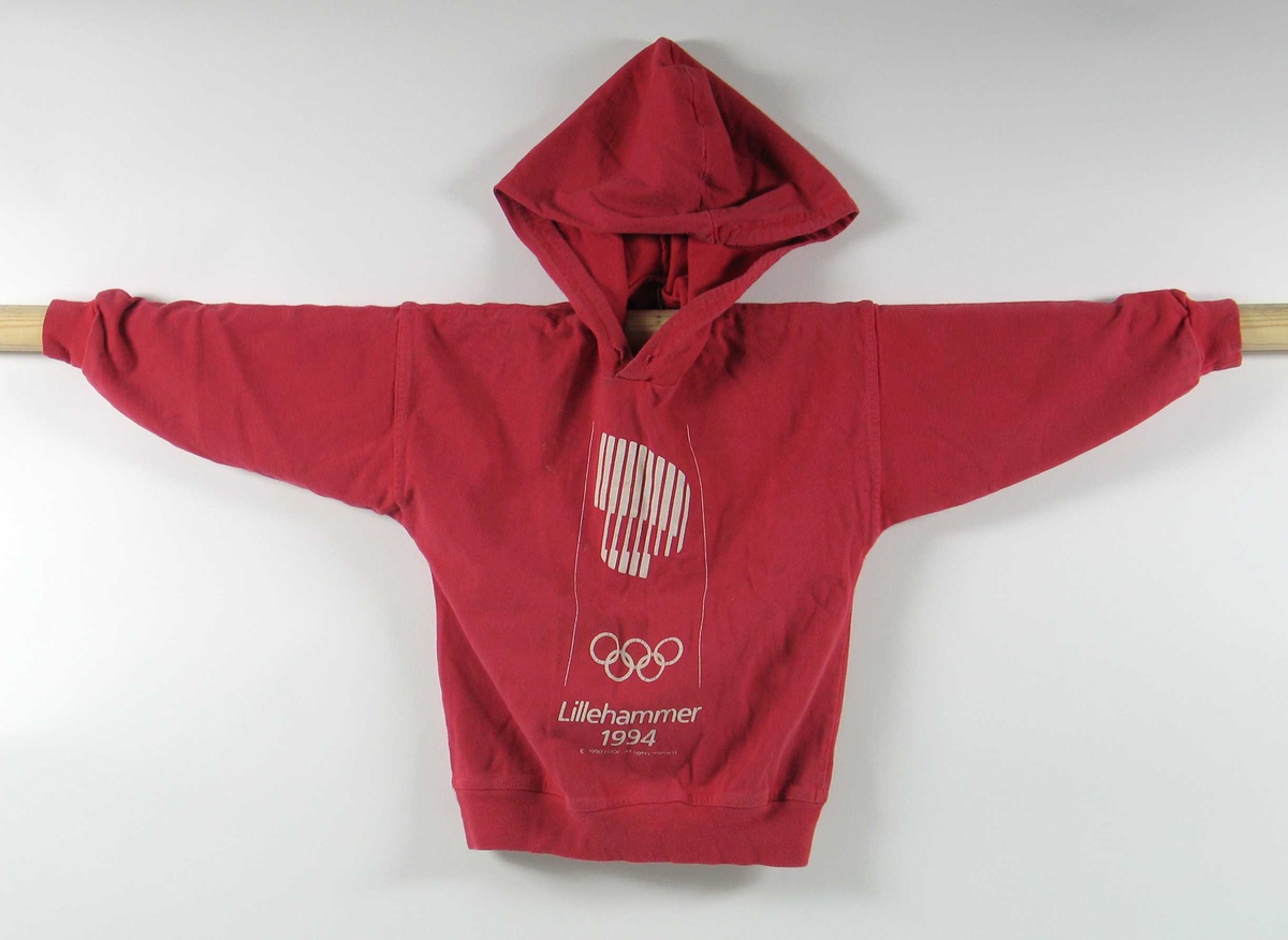 Rød hettegenser i barnestørrelse. Foran er det en logo for de olympiske vinterleker på Lillehammer i 1994. Hull til hettestramming i halsen, men snoren mangler.