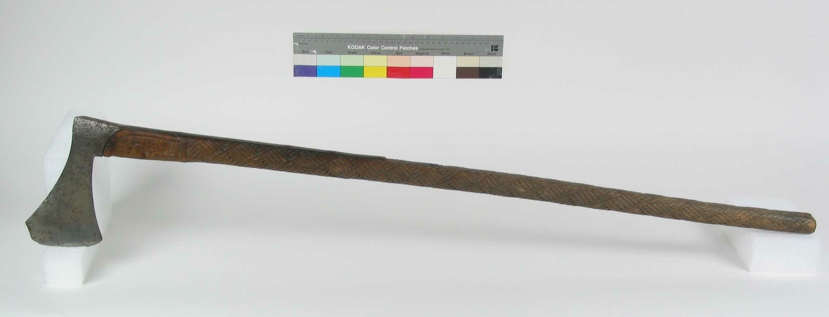 Asymmetrisk økseblad, sammensveiset med overliggende jernskinne.
Fremre øksebladspiss brukket.
Rikt dekorert skaft med flettet rutemotiv. Innskripsjon på øvre del av skaftet (se Påført tekst)
