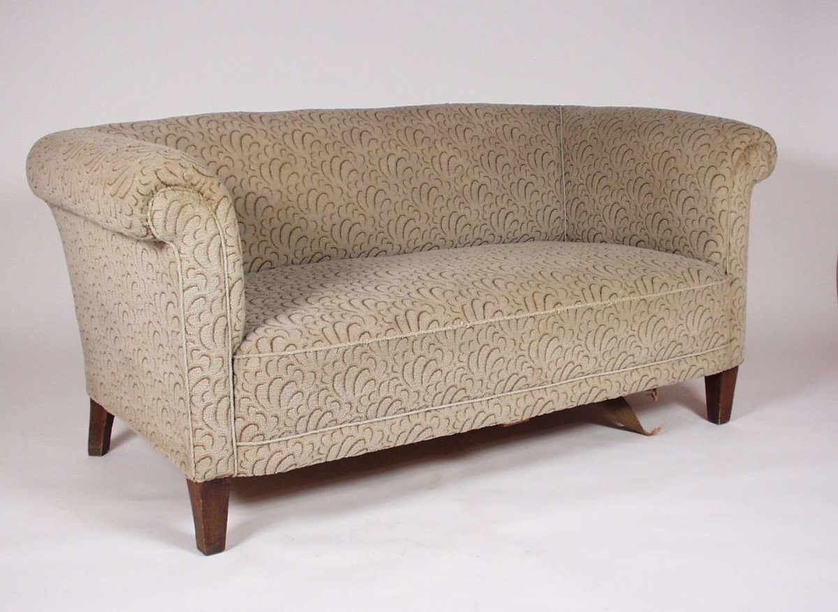 Sofaen har ramme av tre, og er stoppet med mønstret brunt og beige tekstil. Et jutebånd under sofaen er løsnet.