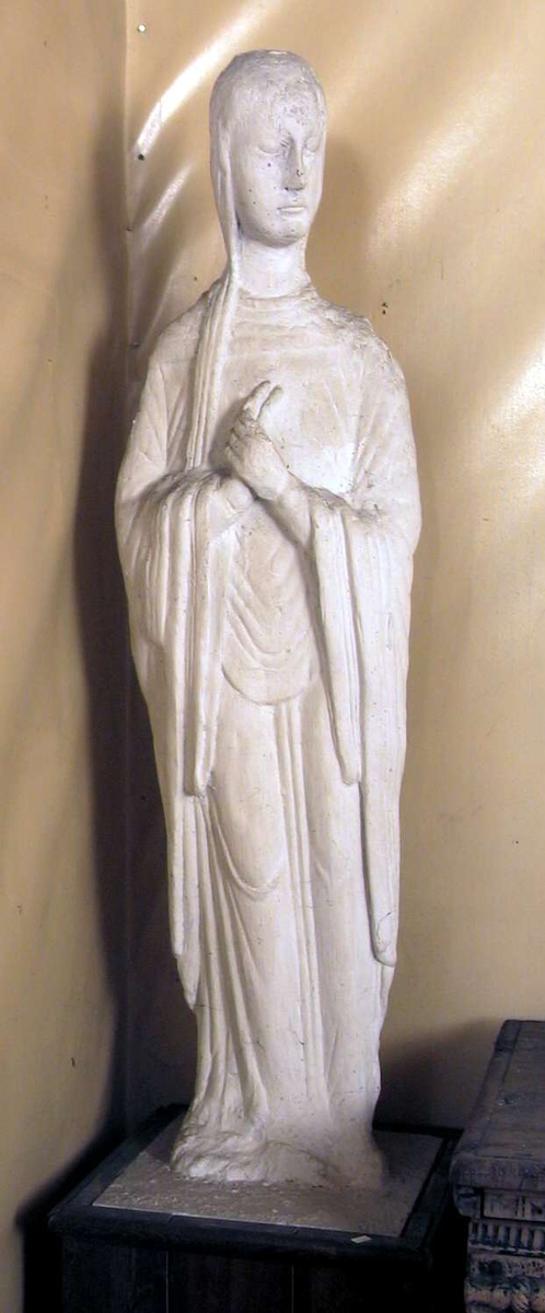 En avstøpning i gips av en treskulptur av jomfru Maria. Skulpturen sår på en sokkel kledd med tre.