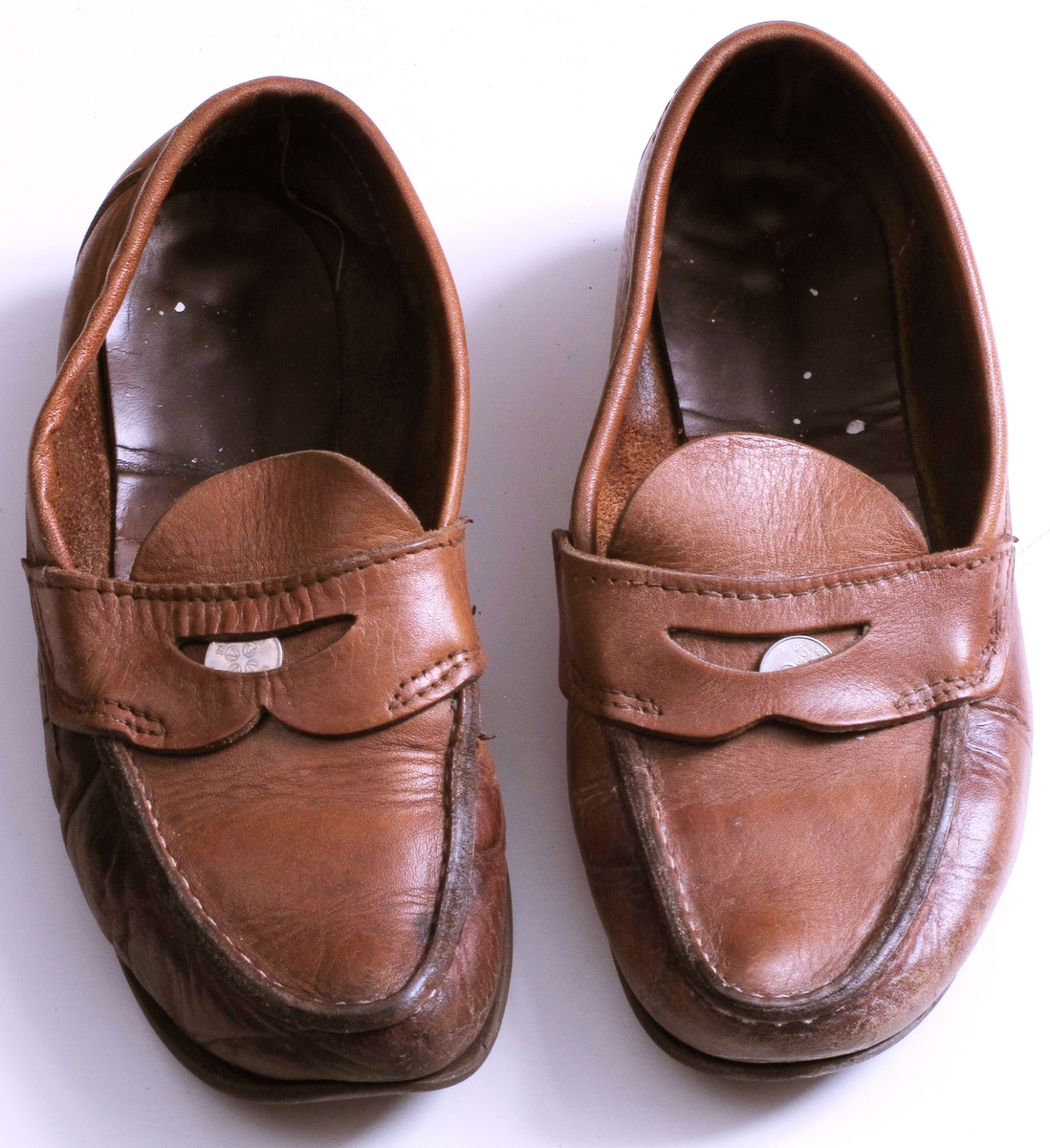 Brune makkasiner i lær, med brune gummisåler.  Lomme foran med 25-ører i.  Høyre sko har en metallstift i sålen rett foran hælen.  Velbrukte, høyre sko er gjennomslitt på utsiden av foten, og med malingflekker oppi.