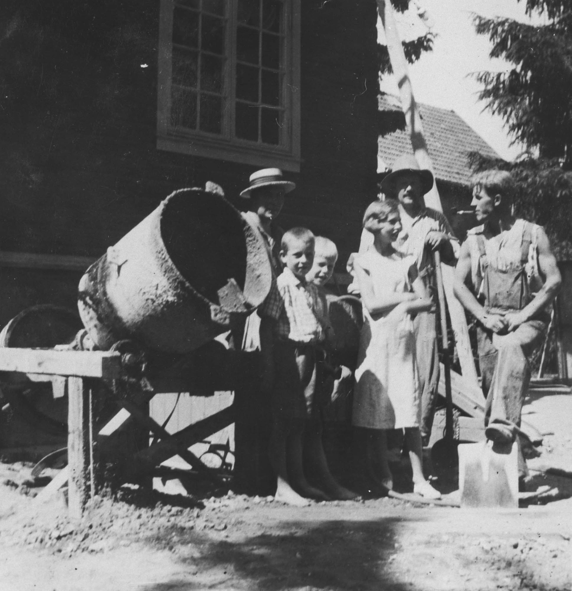 Tre voksne og tre barn ved en sementblander.Bildet er tatt foran kjemi- eller fabrikkbygning.