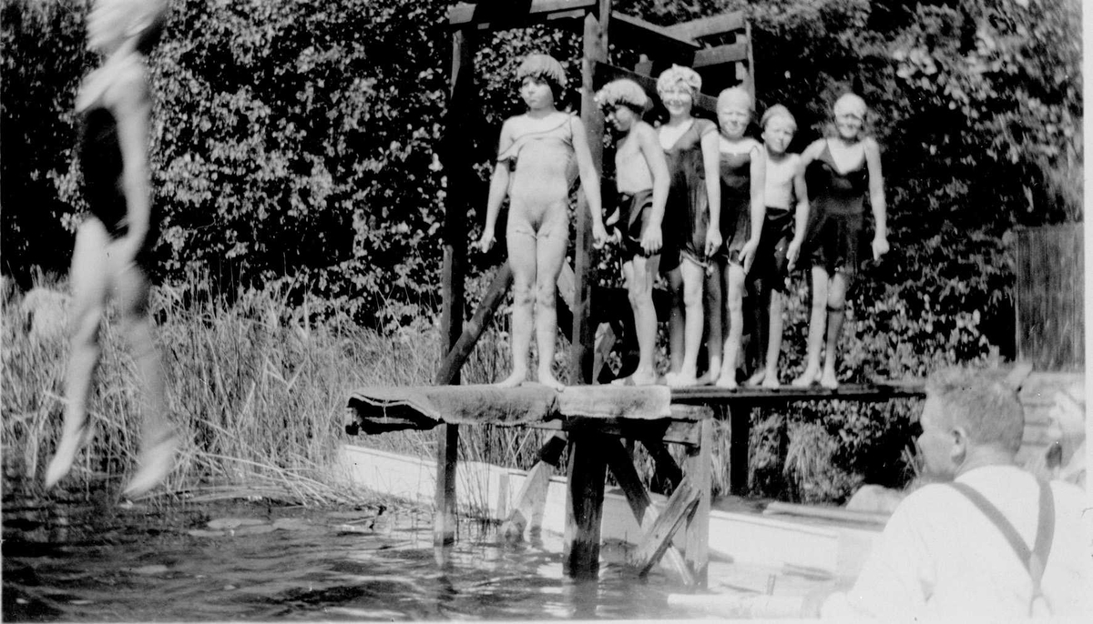 Bading i Langtjernet, østsiden ved Vangen. Barn iført badedrakter og badehetter hopper fra stypebrettet. En voksen mann er tilskuer.