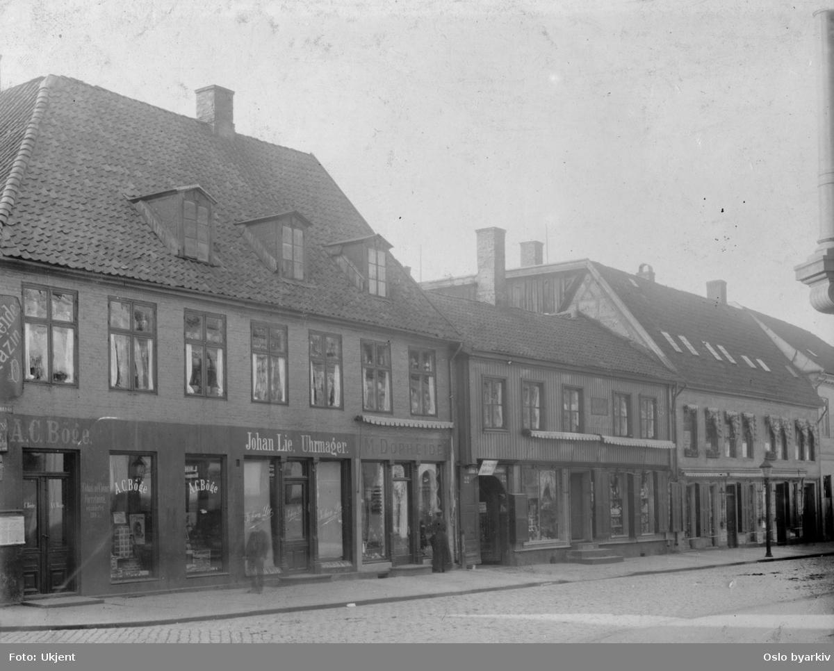 Gatemiljø med butikker. Johan Lie, Uhrmager, Storgata 16. Stadsingeniøren - Fotografier af Kristiania (albumtittel). Ca 1892, basert på øvrige daterte bilder i samme album.