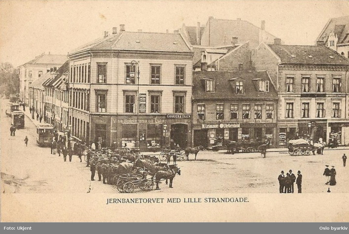 Jernbanetorget ved Lille Strandgate (Strandgata fra 1934) til venstre. Hestedrosjer oppstilt, hestetransport, butikker, Blåtrikker. Gamle bygårder med butikker der Amerikalinjens bygning står idag. Postkort.
