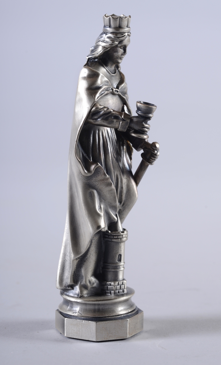 Blankpolert statuett av St. Barbara, skytshelgen for blant annet gruvearbeidere. Hun har en krone på hodet, holder en kopp og et sverd, og står over en steinbygning.