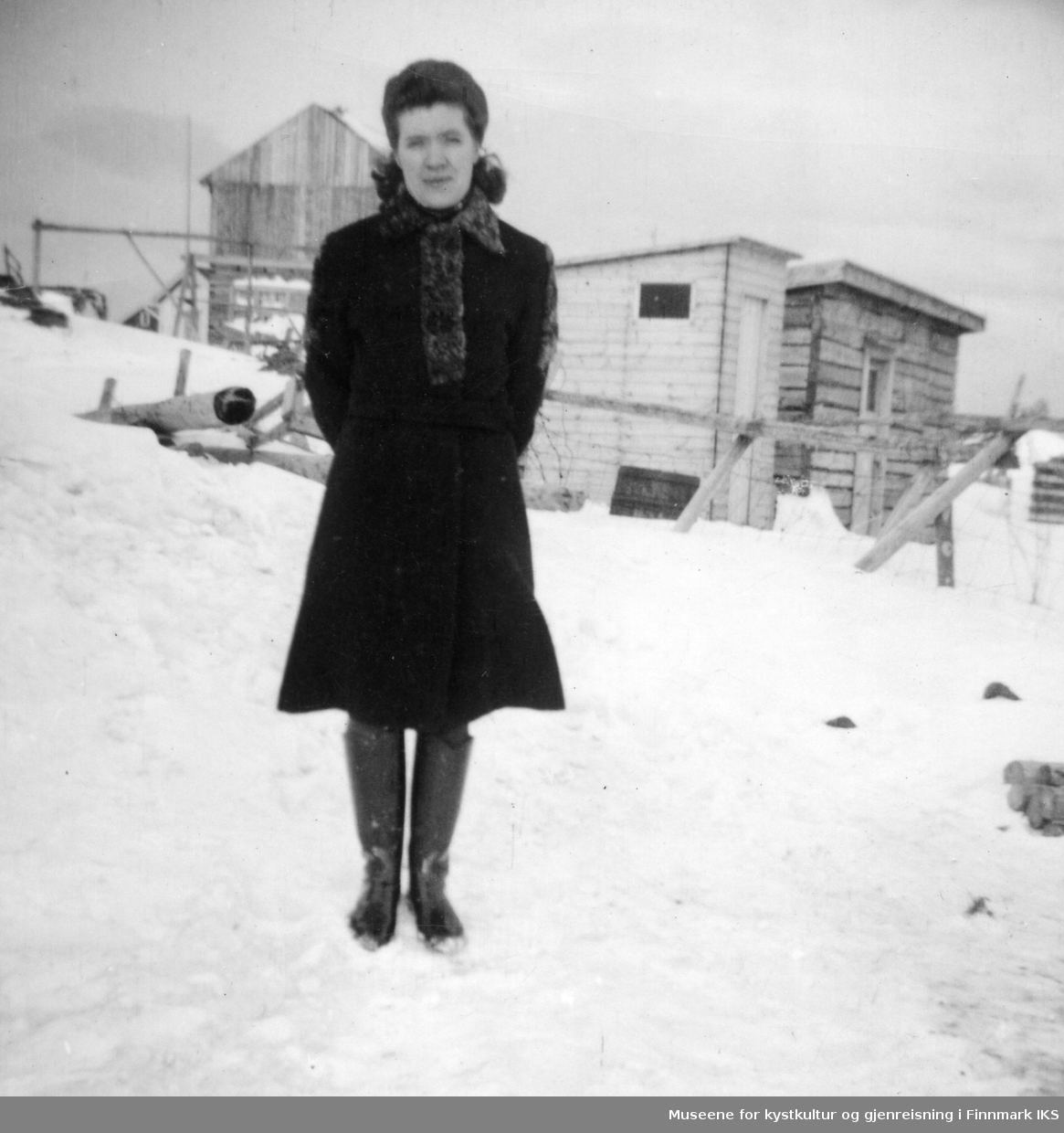 Rikarda Kristensen i kåpe og støvler. Arthur Ingebrigtsens hus i bakgrunnen.
Midt på bildet et utedo, ca 1954.