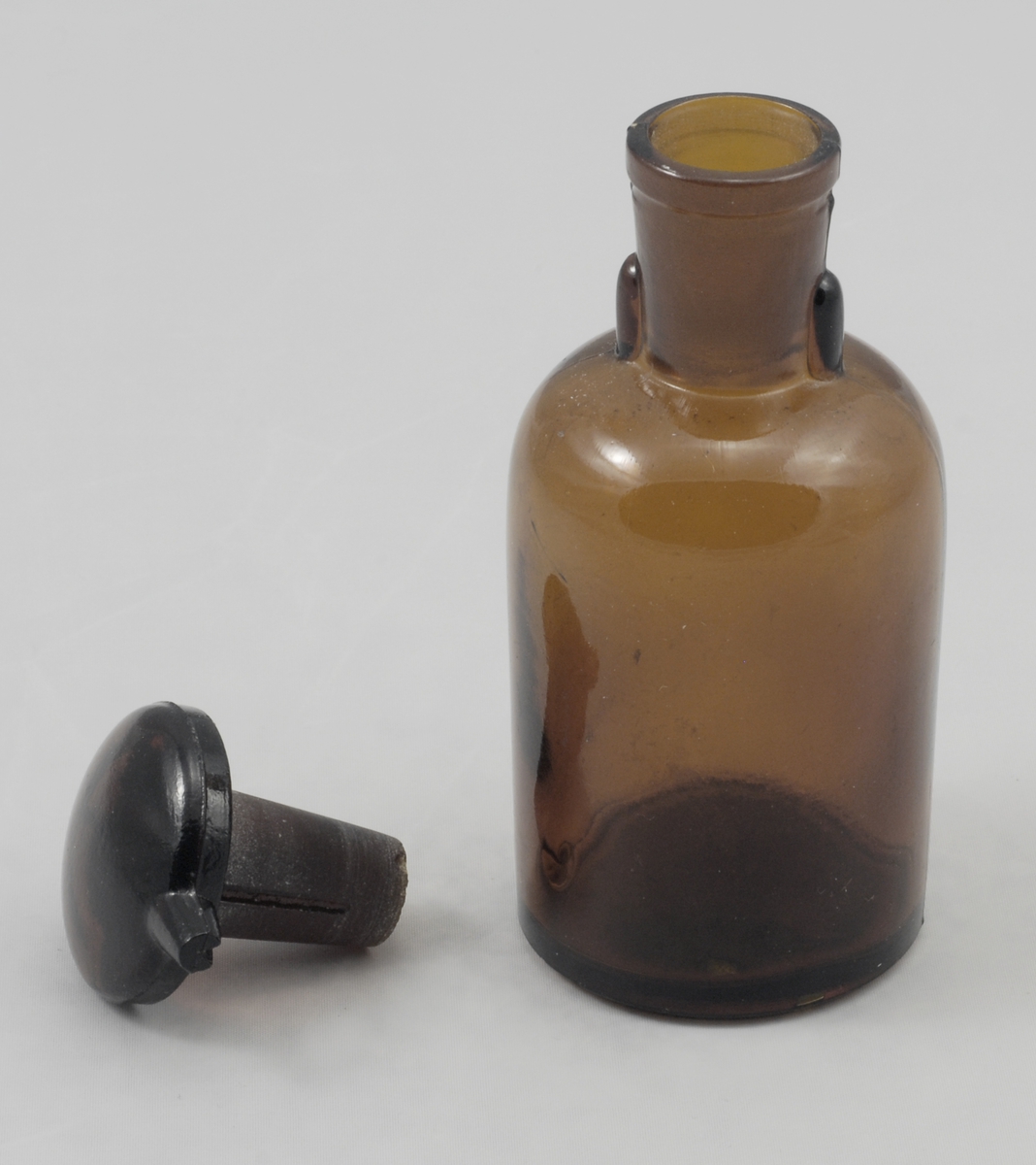 Sylindrisk brun glassflaske med markert avrundet skulder, sylindrisk, lettere konisk hals. På toppen en kork med soppform, korken har rift i stammen og hatten til uthelling av væske.