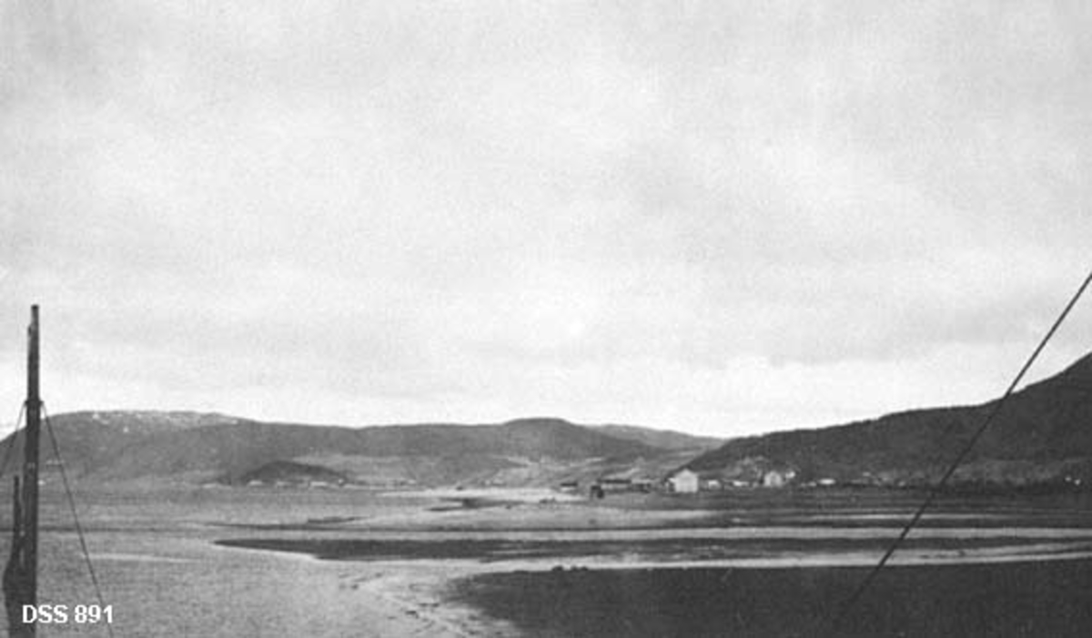 Utsikt fra Mosjøen mot Halsøy.  I forgrunnen ses noe som kan minne om barduntau mot ei elveør.  Op høyre side av fjorden ses en del bebyggelse på sletta mot vatnet.  Bakenfor ligger det skogkledde åser. 