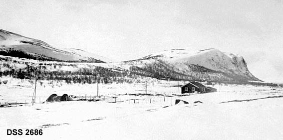 Utsikt fra jernbanelinja på Dovrefjell, Anfinnsbru og Vålåsjøkampen i bakgrunnen.  Snødekt høgfjellslandskap med hytte, fjellformasjon med fjellbjørk rundt «foten» i bakgrunnen.
