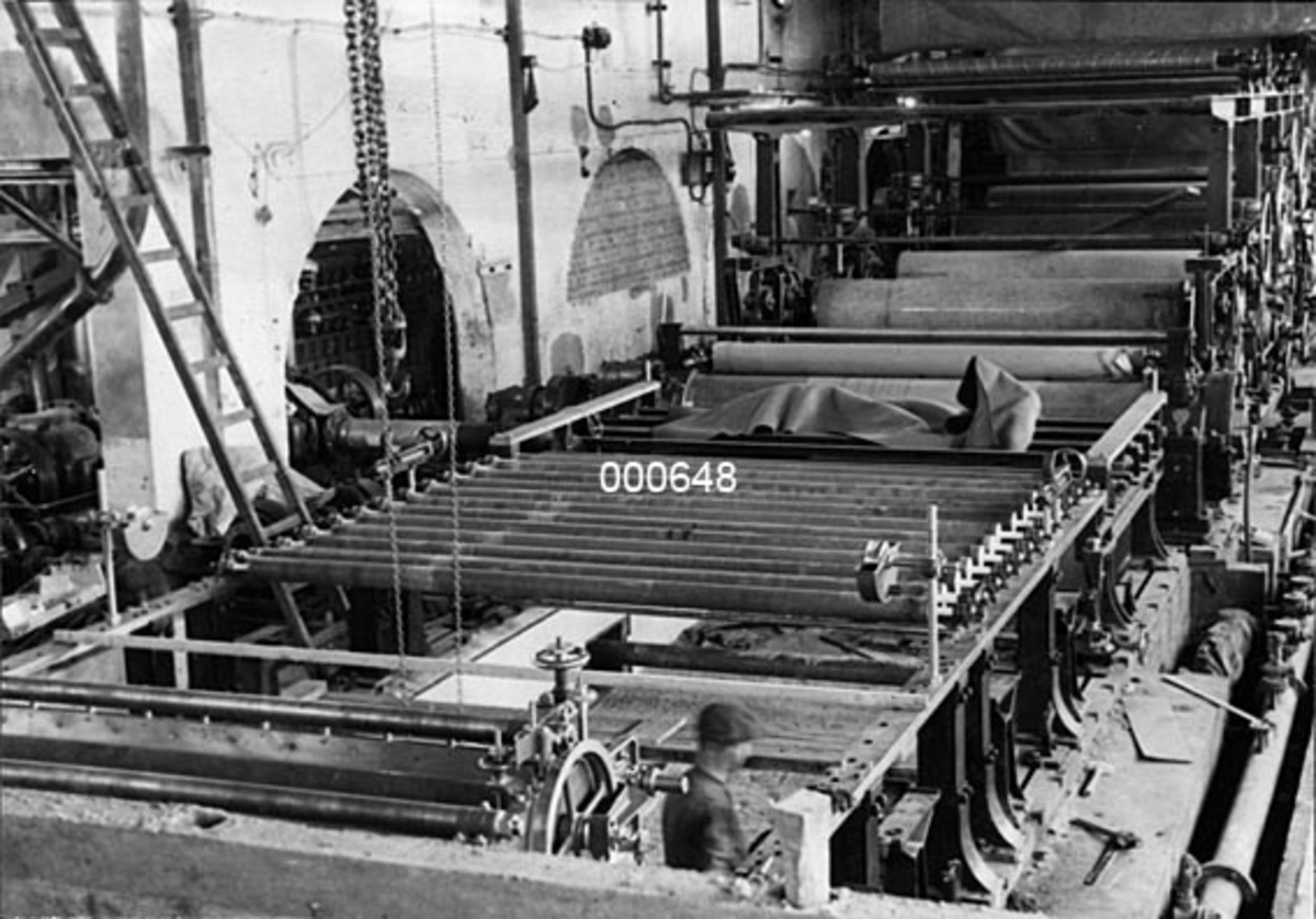 Interiør fra maskinsalen ved A/S Buskerud Papirfabrikk på Åssida i Drammen (den gang i Lier kommune).  Fotografiet er tatt under ombygging av papirmaskinen i 1937.  Det er tatt fra et høyt punkt i enden av maskinsalen, antakelig fra toppen av massekummen.  Det viser nytt vire- og pressparti under montering.  Tørkepartiet i bakgrunnen er dekt av en presenning.  Ombygginga ble gjort med sikte på å øke driftshastigheten fra 4600 tonn i året til 6500-7000 tonn per år,  For å kunne gjøre det var det nødvendig å fornye så vel papirmaskinen som driftsmaskineriet.  I første omgang var det vire- og presspartiet som ble ombygd og fikk elektrisk drift.  Virepartiet ble også forlenget med cirka tre meter slik at lengden ble 21 300 millimeter.  Til venstre i forgrunnen skimtes brystvalsen med rystepapparat, som er i ferd med å bli heist på plass.  Også de bakerste registervalsene er ferdigmonterte.  I bakgrunnen ses presspartiet og det presenningdekte tørkepartiet. 