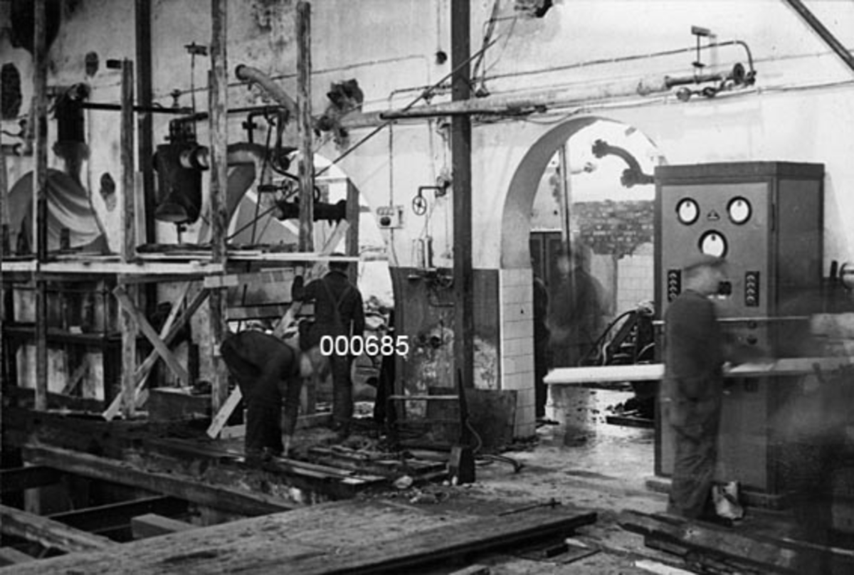Interiør fra A/S Buskerud Papirfabrikk på Åssida i Drammen (den gang i Lier kommune).  I 1937 ble papirmaskinen ved denne fabrikken fullstendig ombygd, særlig for å kunne kjøre maskinen med større driftshastigheter og dermed oppnå større produksjonsvolumer.  Fotografiet er tatt under ombyggingsarbeidet.  Det viser den delen av maskinsalen hvor papirmaskinens tørkeparti har stått.  Da fotografiet ble tatt var denne delen av maskinen demontert og en diger lem var lagt over maskingrava for å gi muligheter for passasje.  Langs veggen mot det som til da hadde vært transmisjonsrom hadde arbeiderne satt opp et trestillas.  Vi ser også inn gjennom en rundbuet døråpning mot dette rommet.  Til høyre for døråpningen står det en mann foran et elektrisk skap.  Dette ble levert av Siemens i forbindelse med ombyggingsprosessen.  

Omlegging fra dampdrift til elektrisk energi preget mange papirfabrikker på denne tida.  I 1937 reklamerte Siemens i Papir-Journalen med at de hadde levert elektroviklere til Svelvik Papirfabrik, Brager Papirfabrik, Buskerud Papirfabrik, Saugbrugsforeningen (3 stk. ), Jarlsberg Paper Mills (2 stk. ) og Star Paper Mill.  Også Granfos Brug hadde bestilt slikt materiell. 