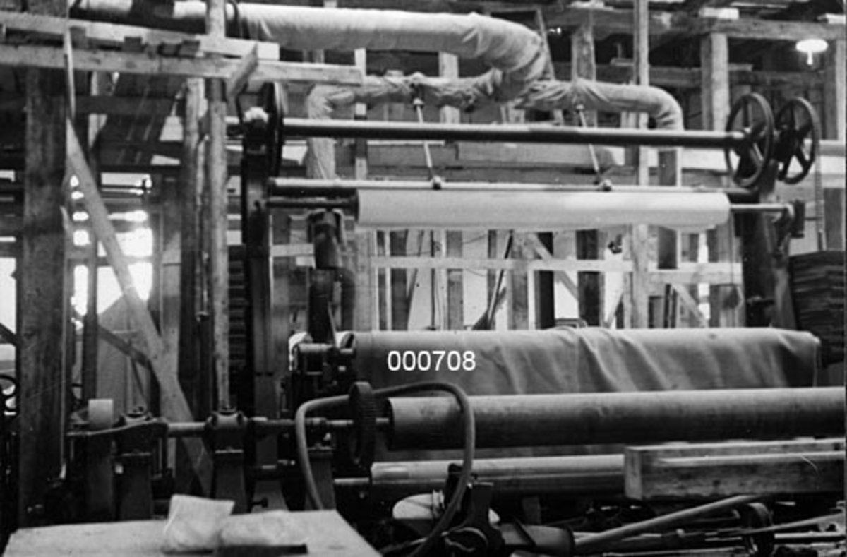 Interiør fra A/S Buskerud Papirfabrikk på Åssida i Drammen (den gang i Lier kommune).  I 1937 ble papirmaskinen ved denne fabrikken fullstendig ombygd, særlig for å kunne kjøre maskinen med større driftshastigheter slik at en kunne oppnå større produksjonsvolumer.  I den forbindelse la bedriften om fra dampdrift til elektrisk drift.  Fotografiet er tatt fra den indre enden av maskinsalen, mot en ny vegg som ble satt opp mot den tilstøtende kuttersalen.  Veggen ble utført i bindingsverk, og da fotografiet ble tatt var det bare skjellettkonstruksjonen som var oppsatt, uten kledning.  I forgrunnen står en del maskiner, som antakelig bare er satt til side i forbindelse med ombyggingsarbeidene. 