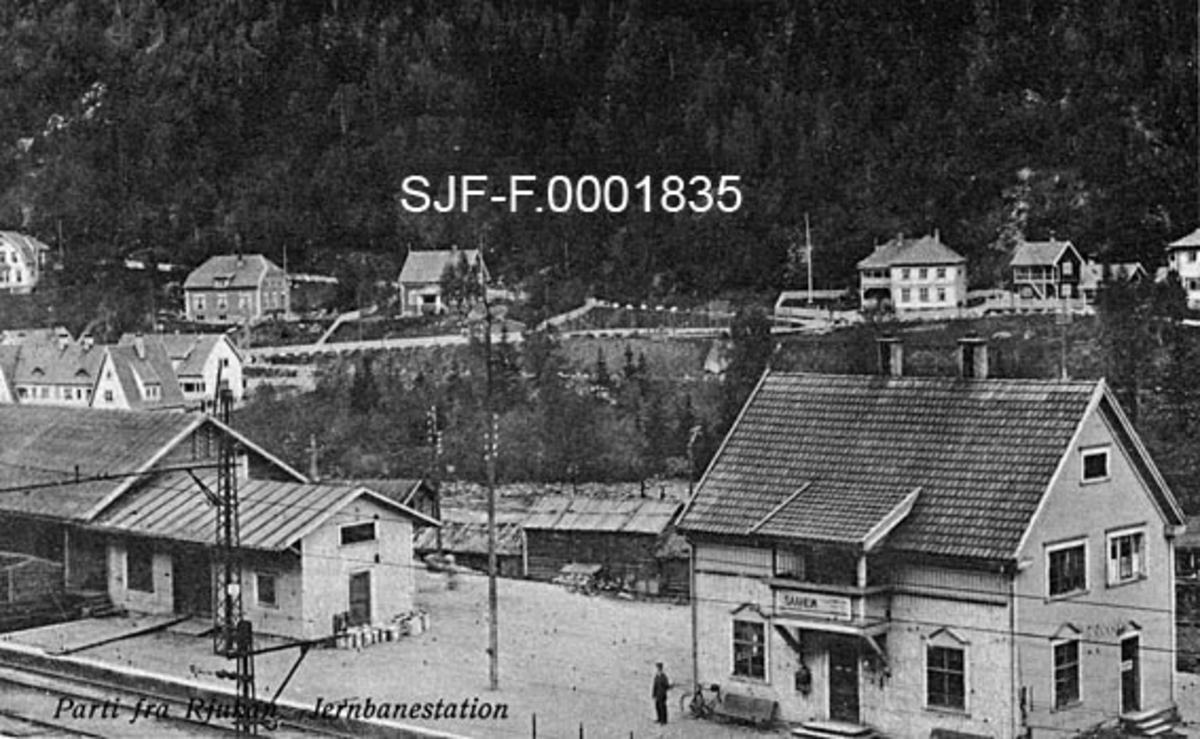 Rjukan jernbanestasjon i Tinn i Telemark.  Rjukan, eller Såheim som stedet en gang ble kalt, ble utbygd tidlig på 1900-tallet, da Sam Eyde på vegne av Norsk Hydro kjøpte fallrettigheter i elva Måna i Vestfjorddalen, for å kunne videreutvikle en industriell virksomhet som allerede var i god gang på Notodden. Et viktig ledd i denne satsinga var jernbaneforbindelsen mellom de to stedene, Rjukanbanen eller Vestfjorddalsbanen, som den opprinnelig ble kalt.  Denne banestrekningen ble åpnet i 1909 og var i drift til 1991.  På den 30 kilometer lange strekningen over Tinnsjøen gikk jernbanevognene på ferje.  Rjukanbanen var ellers 
Norges første normalsporede elektriske jernbane, noe som illustreres av ledninger i forgrunnen på dette fotografiet.  Statsjonsbygningen er et toetasjes trehus med saltak.  Den har dør midt på langveggen ut mot perrongen, og en veranda i annen etasje over denne døra.  På verandaveggen er skiltet med stasjonens navn - Såheim - og høyde over havet påsatt.  Til venstre på bildet ser vi stasjonens pakk- og godshus.  Mellom stasjonsbygningene, nærmere elva, skimter vi ei uthusrekke fra et gardstun som sto her før Rjukan ble industristed.  På motsatt side av elva ligger en del større trevillaer, antakelig bygd for funksjonærskiktet i det nye samfunnet.  