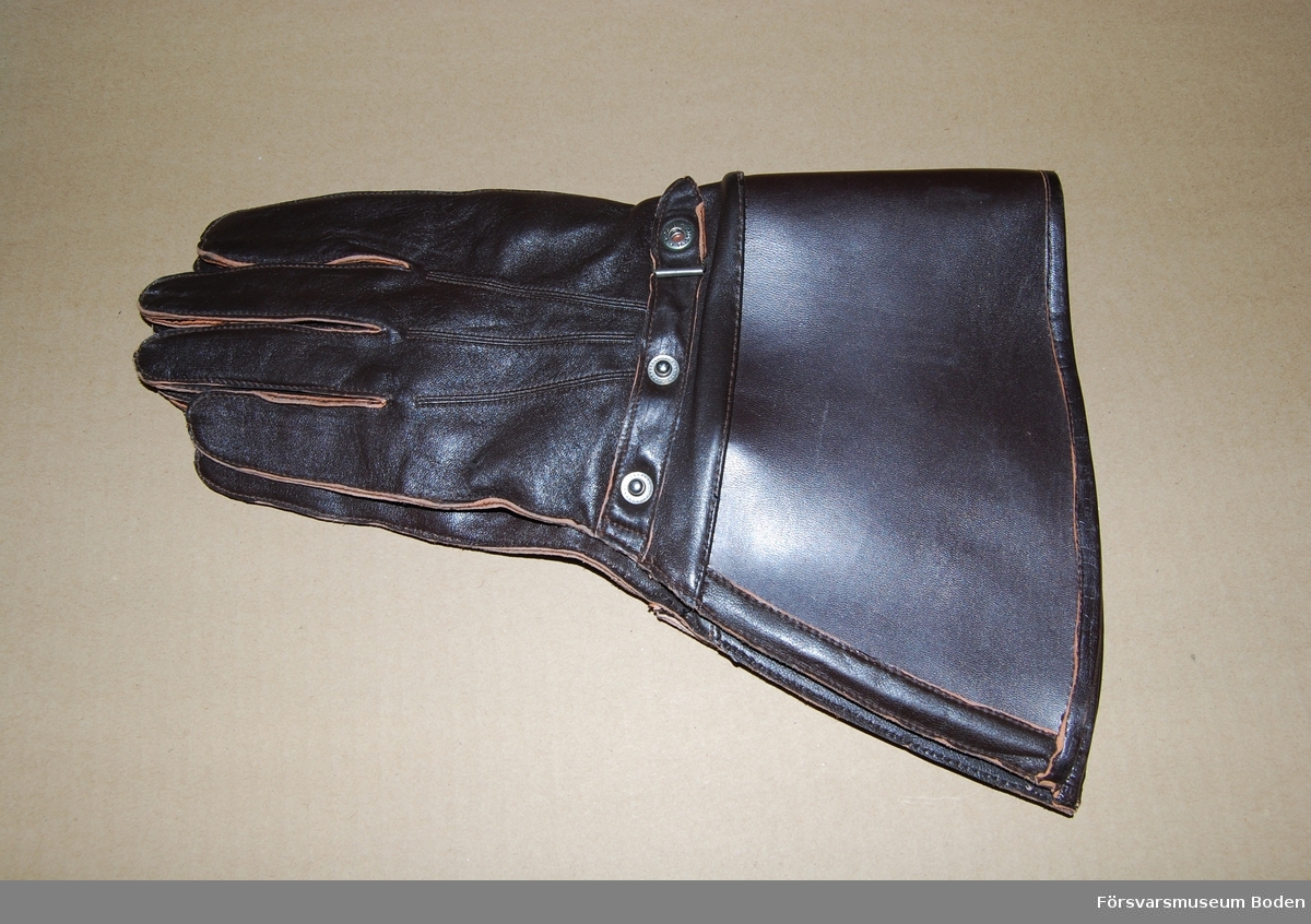 Ett par oanvända handskar av mörkbrunt läder med styv krage som är präglad med tre kronor vid kanten på utsidan. Spänntamp med tryckknapp på ovansidan. Handskarna är hopfästa med tråd. Avsedd att användas av bl.a. mc-ordonnanser.