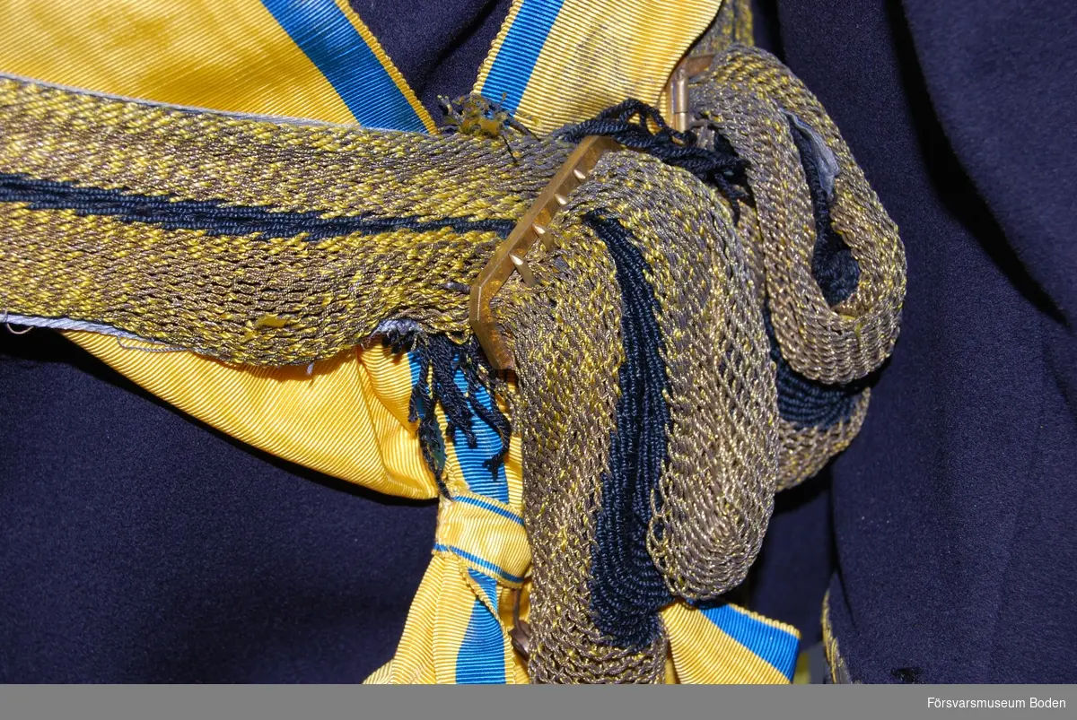 Virkat skärp i guldfärgat silke med blå rand i centrum. Två tofsar av blått silke med tjockare guldfrans  ytterst. Spänne av mässing för justering av längden.
