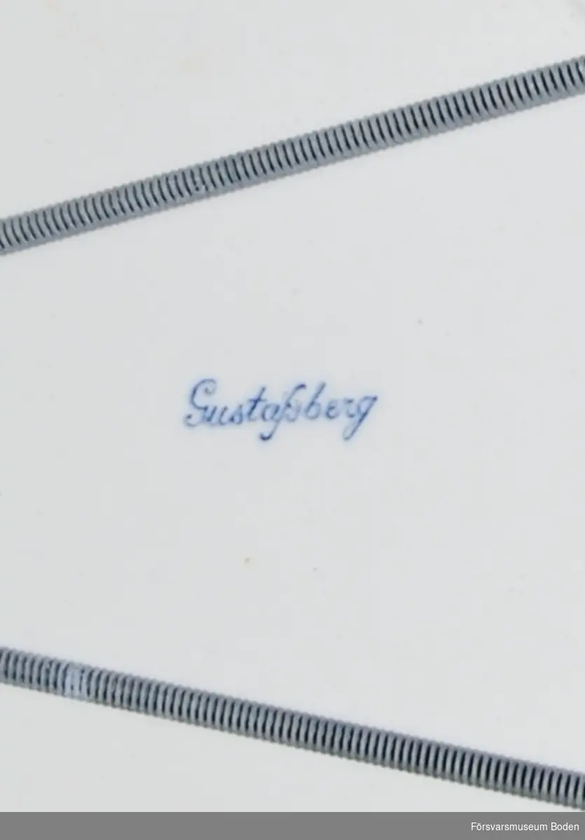 Vitt porslin av Gustavsbergs tillverkning med krona och bokstäverna BF. Tallrikshållare för upphängning monterad.