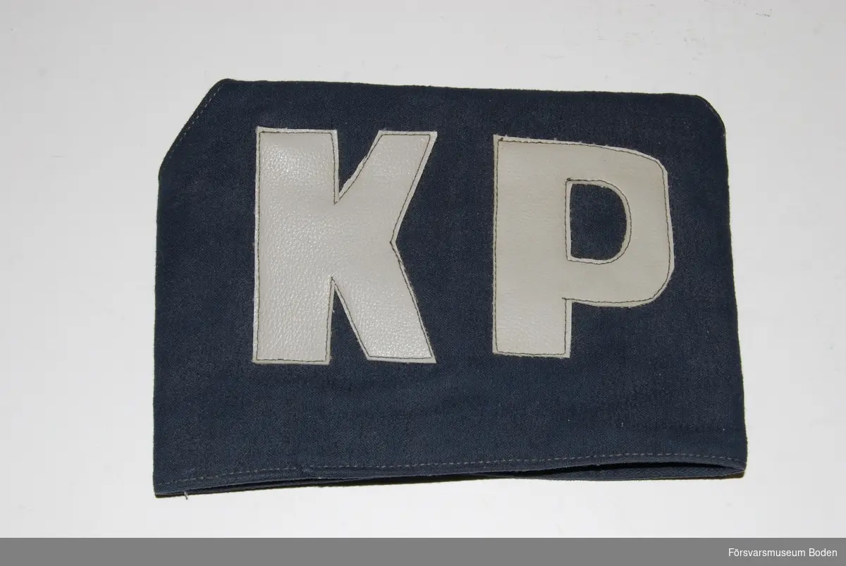 Bindel av mörkblått tyg, troligen handsydd och försedd med bokstäverna "KP" i grå vinylplast. Kardborrelåsning på baksidan.