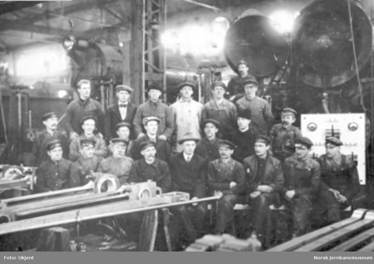 En gruppe verkstedarbeidere oppstilt for fotografering med damplokomotiver i bakgrunnen