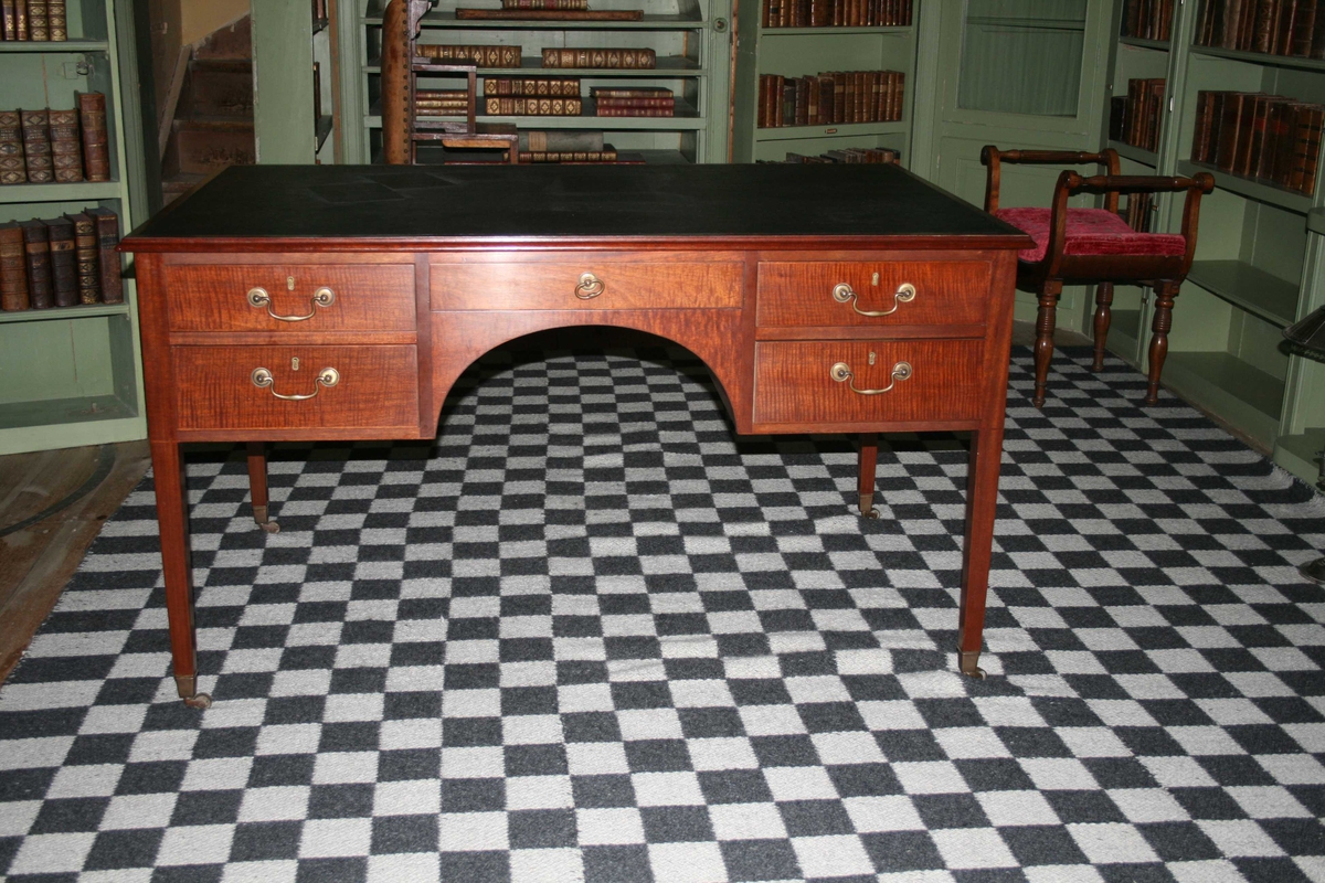 Skrivebord i mahogni, bordplate trukket med svart kunstlær. Fem skuffer på hver side av bordet. Beslag og trinser i messing.