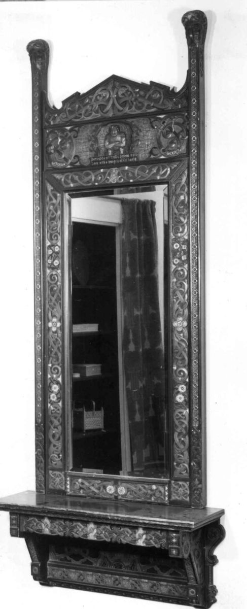 Rektangulært, høyreist speil med flerfarget utskåret og malt ornamentikk i form av blomster, akantus og voluttranker. Sentrert i øvre del av speil er et utskåret troll med innskrift. I hvert øvre hjørne står det opp et utskåret hode. Til speilet hører et konsollbord.