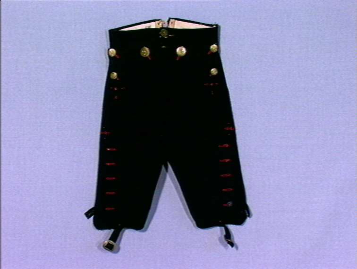 Svart bukse, knapphull er sydd med rød tråd.
