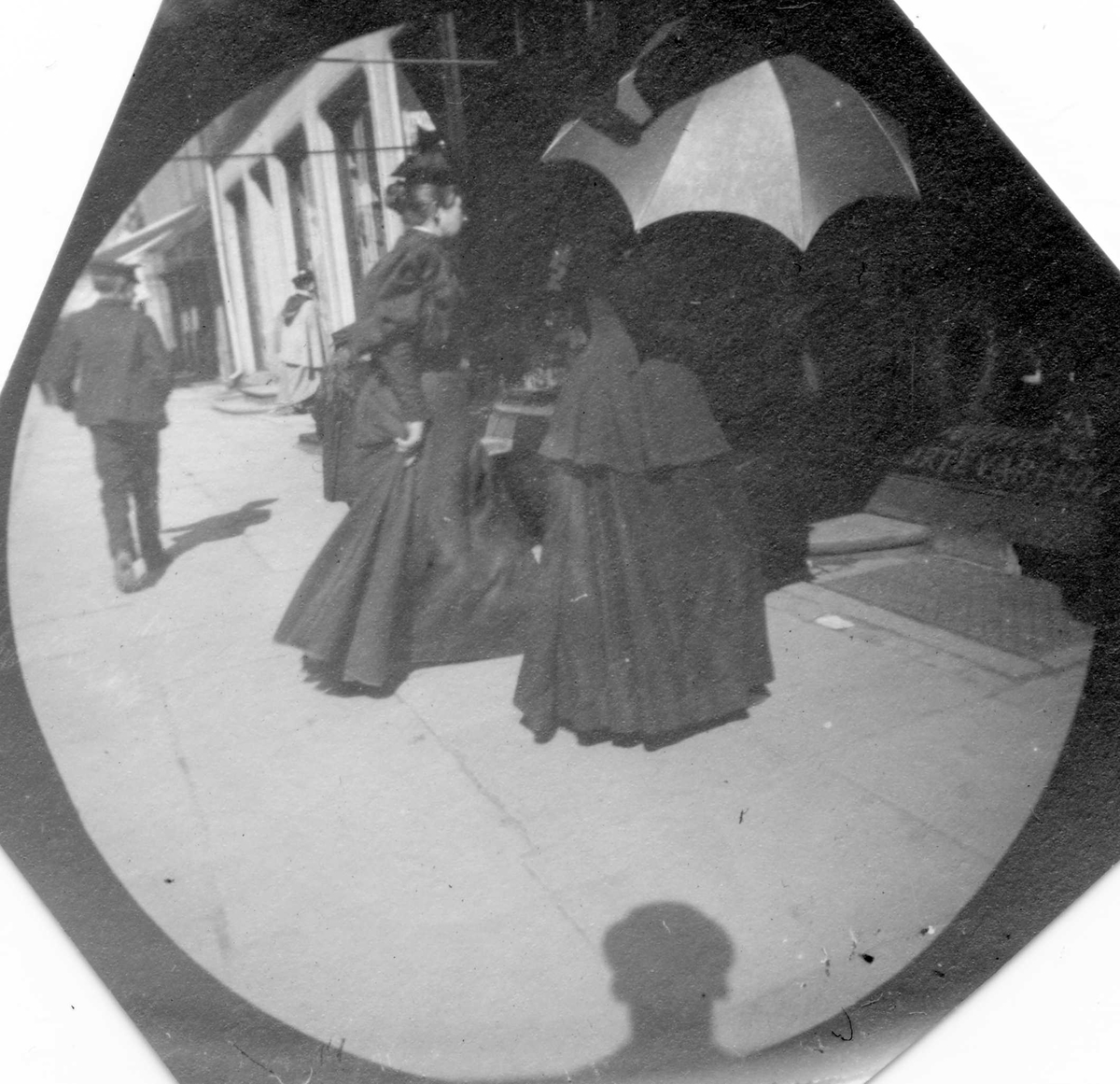 To kvinner, en med lys paraply og en mann i bygate, ant. Oslo. Butikker.