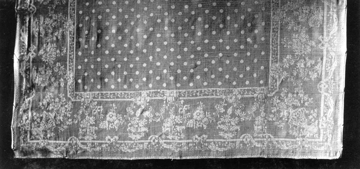 Hvit damaskserviett med prikket bunn. Bred kant med striper, blomster og lamberquins. Antagelig fra Tyskland slutten av 1700-tallet.