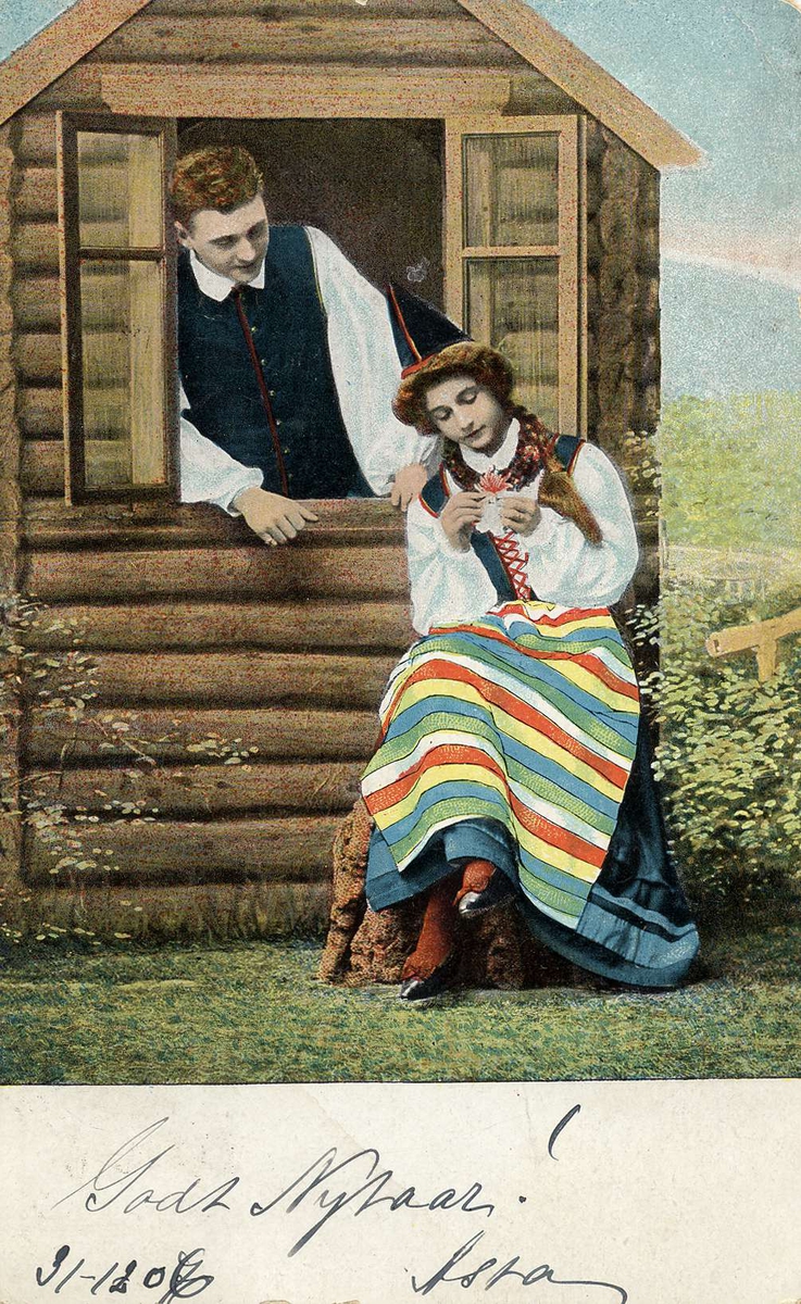 Postkort. Nyttårshilsen. En kvinne i folkedrakt sitter utenfor ei lita hytte. Hun sitter med en blomst i hånden. I vindusåpningen på hytta står en mann og ser på henne. Poststemplet 31.12.1906