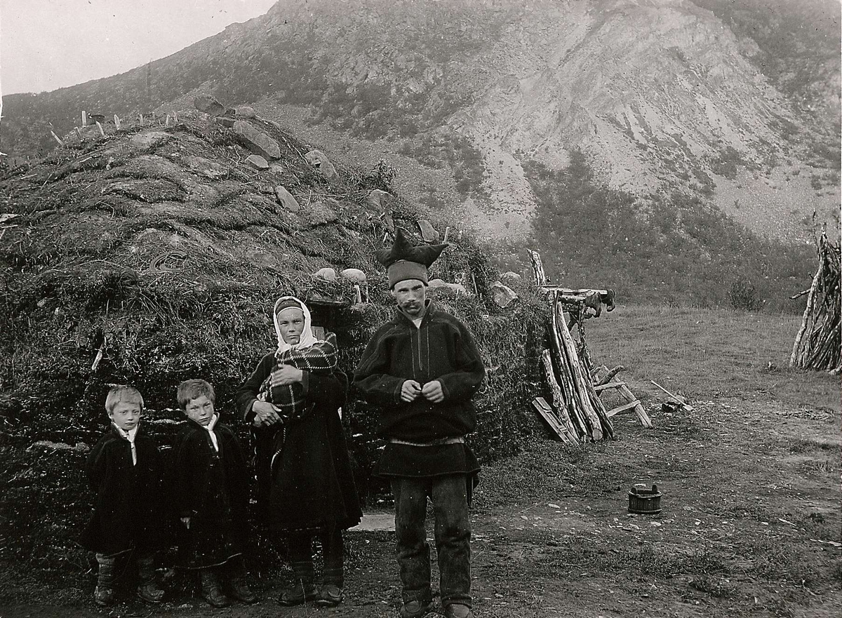 Familiegruppe (5) i samisk drakt, oppstilt foran gamme ved Sjilsjokka (Suossjohka?) i Laksefjord, Lebesby. Kvinnen holder et spebarn, mens sønnene Erik og Ole står ved siden av.
Del av serie fra en forskningsreise i Øst-Finnmark 1909.
