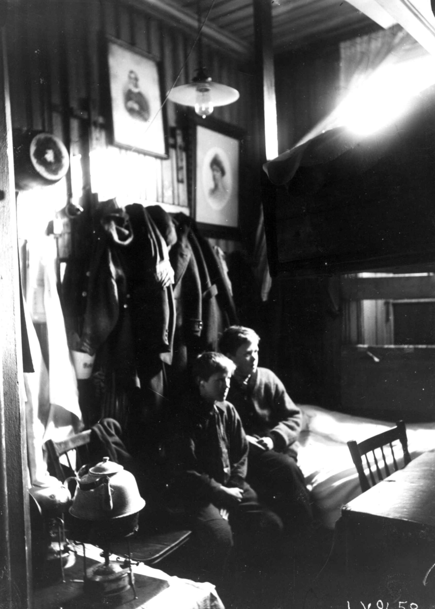 Interiør, Grønlandsstrøket, Oslo. Bebodd bakrom i butikk, 2 gutter sitter mellom seng og kjøkkenkrok med kokeapparat i tett møblert rom.
Fra boliginspektør Nanna Brochs boligundersøkelser i Oslo 1920-årene.
Se Relaterte objekter.