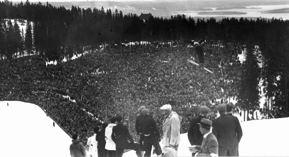 Ski. Hopprenn. Holmenkollen, Oslo 1934. Oversiktsbilde fra toppen av bakken ned mot
sletta og publikum. Hopper i svevet. Tilskuere ses øverst i unnarennet. Oslofjorden skimtes i bakgrunnen.
