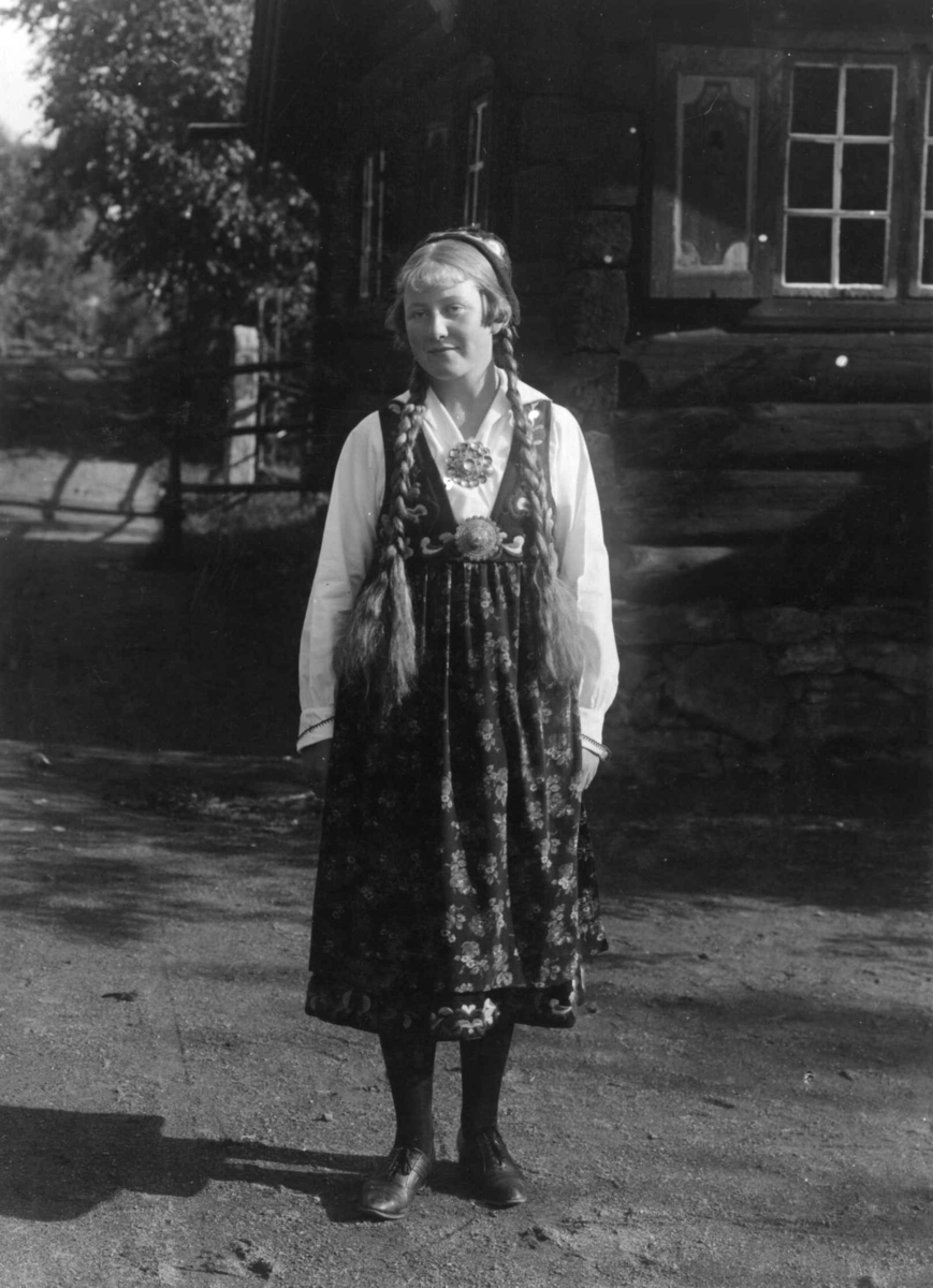 Vert, antagelig Gunhild Knudsen, i drakt foran Gulsvikstua, bygning nr. 121 i Hallingdalstunet på Norsk Folkemuseum.