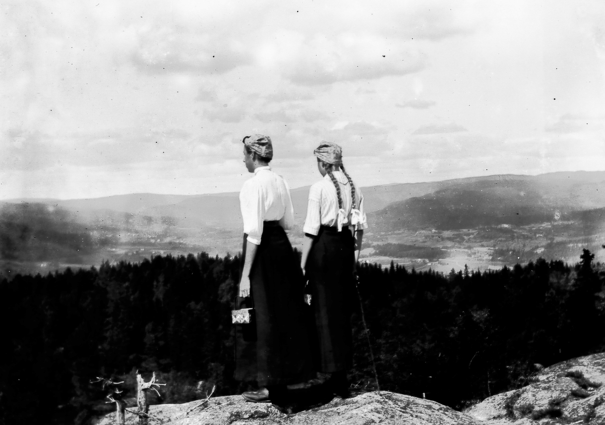 To søstre med sponkurv og hodetørkle ser utover landskapet. Bærplukking?