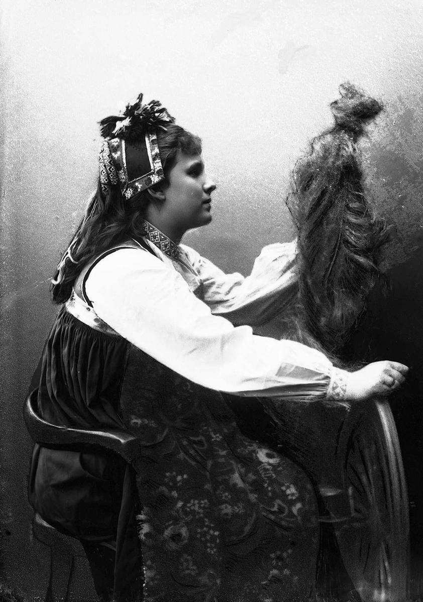 Studioportrett av kvinne i drakt og hodeplagg, som sitter ved en rokk. Bakgrunnen er nøytral.