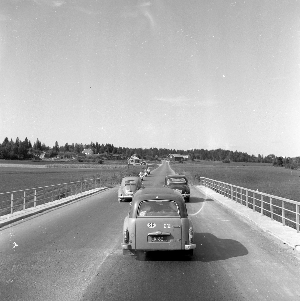 Langs Riksvei 1 kjører tre biler og tre scootere med småhusbebyggelse langs veien. Fotografert 7. juli 1958.