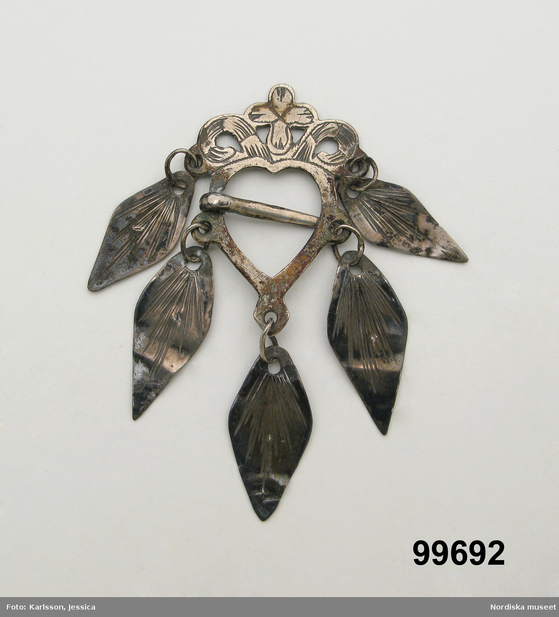 Hjärtformad sölja av silver med ornamenterat krön och 5 lövformade hängen, nål i mitten. Stämplar saknas.

Berit Eldvik maj 2006