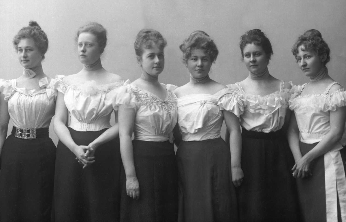 Gruppeportrett, syv kvinner i hvite kortermede bluser og svart skjørt.