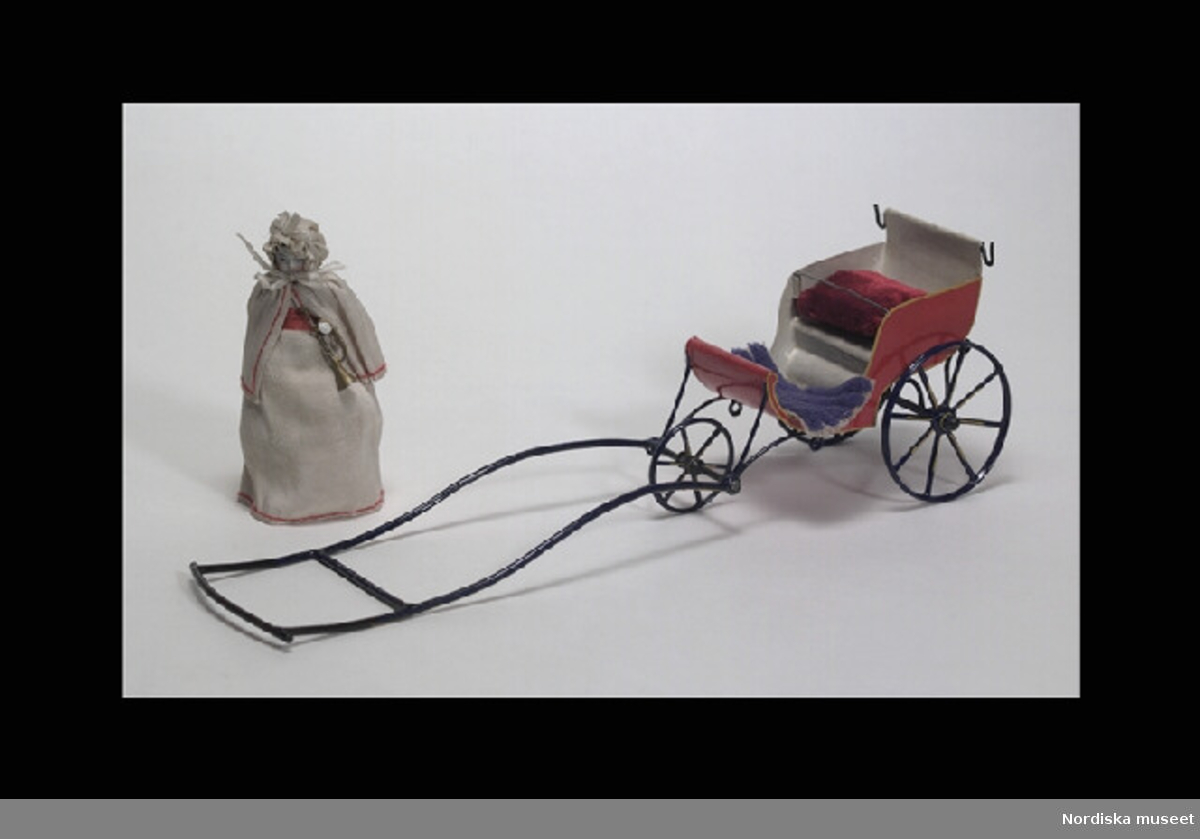 Inventering Sesam 1996-1999:
Vagn   L  32   H  9,7 (cm)  
Docka med klänning  L  11,5 (cm)
Barnvagn/kärra, täcke och kudde (a+) samt docka (b) med trumpet (c), till dockskåp, barnvagn av bockad och rödlackerad plåt, tre blålackerade ekerhjul och långt styre, krokar för sufflett (?), täcke av blå textil, kudde av mörkröd plysch. Docka av vitt porslin, med ena armen böjd, målade ansiktsdrag och svart hår, iklädd vit bärklänning och pelerin, kantsömnad med röd tråd, vitmössa. Trumpet av bronserat tenn.
Tillhör dockskåp från omkr 1840, inv nr 145.505, inrett 1872 av leksakshandlare W A Bandel, Västerlånggatan, Stockholm, för dottern Thyra, vilken vid överlämnandet till museet själv gjort uppställningen av föremålen i dockskåpet. 
Anna Womack dec 1997