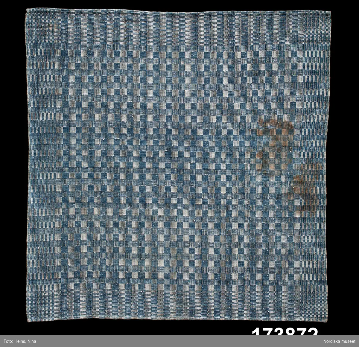 Bordduk.
Varp: 1-trådigt Z-spunnet vitt bomullsgarn, cirka 300 trådar/10 cm.
Inslag: 1-trådigt Z-spunnet blått bomullsgarn.
Förenklad dräll på två partier, så kallad halvdräll. Vävd på fyra skaft. Trädd med 3 trådar i rör enligt väverskan. En duk vävd med bård och schackrutigt mittfält. En smal fåll runt om är sydd på maskin.
/Berit Eldvik 2002