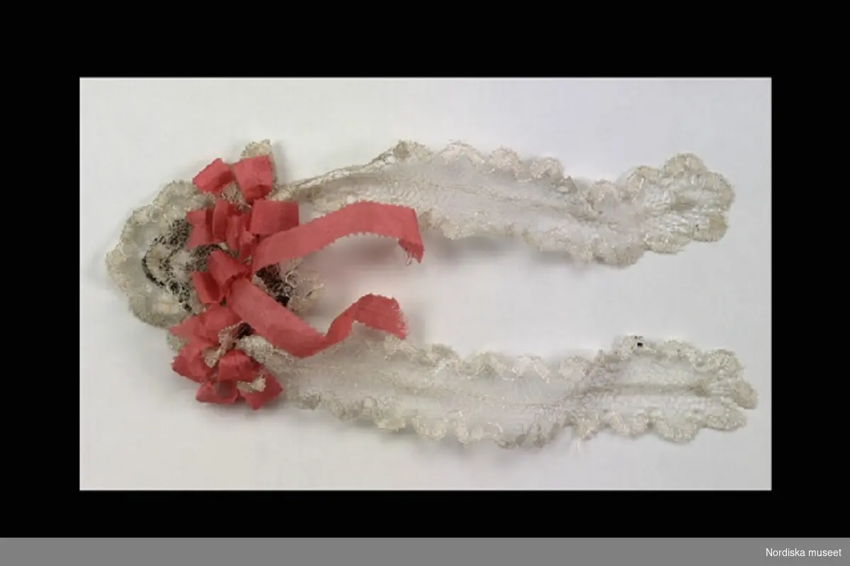 Inventering Sesam 1996-1999:
L 20 cm (med banden)
Dockhårklädsel av vit trekantig spets på brun tyll, utspänd med ståltråd, dekorerad med rosa sidenband och två vita hängande band av spets.
Leif Wallin 1996